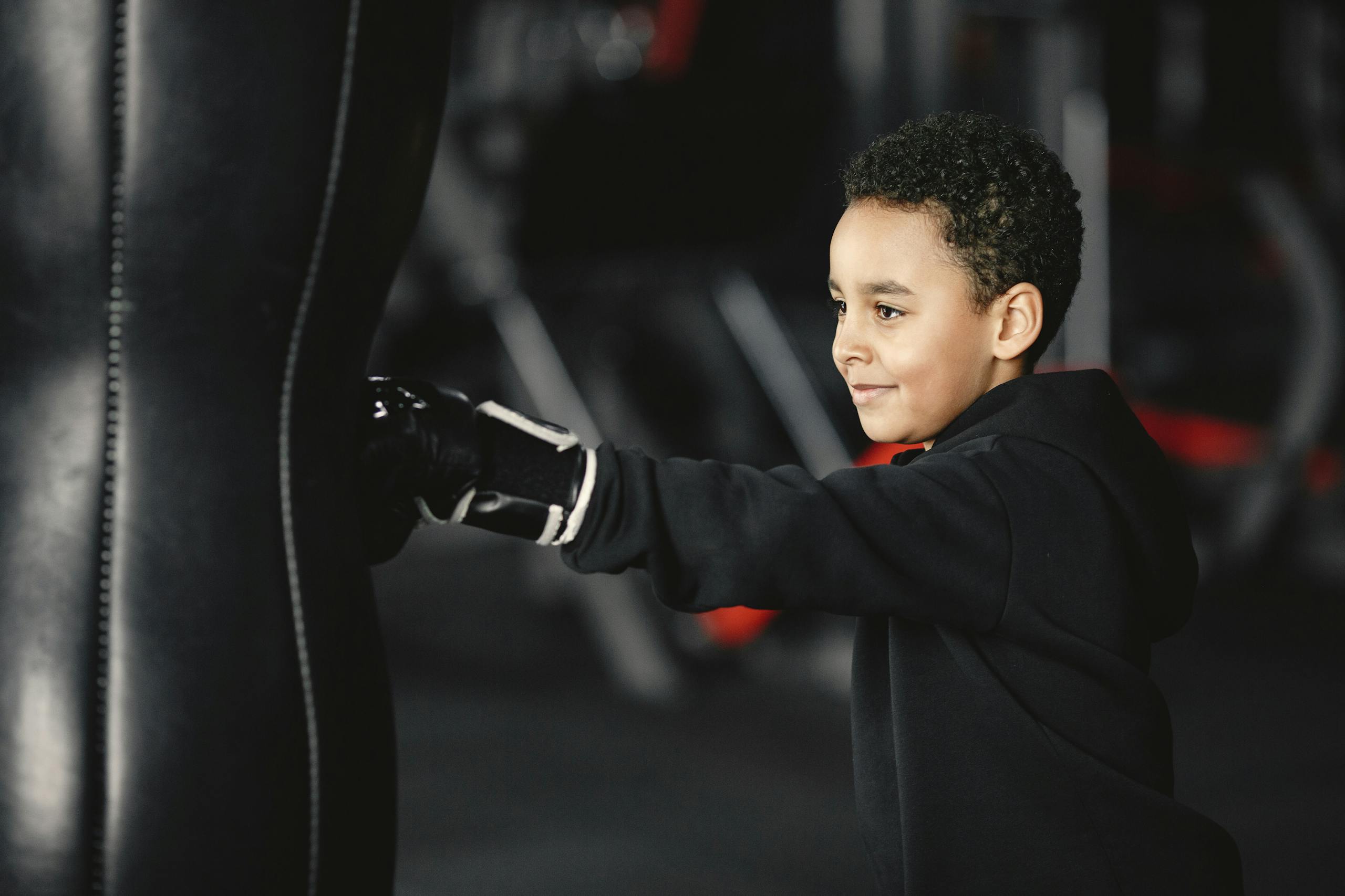 A young boy smiling while training with a punching bag in a gym setting.